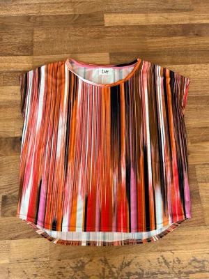 Färgstark randig topp från Isay - Säljer en kortärmad topp från Isay med färgstarka vertikala ränder i orange, svart, rosa, röd och vit. Toppen har rund halsringning och är tillverkad i ett mjukt, lätt material som känns skönt mot huden. 