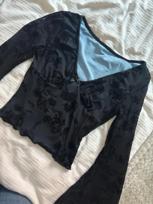 Svart blommig topp med v-ringning - Snygg svart topp med blommigt mönster i sammet och v-ringad hals. Toppen har långa, utsvängda ärmar och croppad passform. Perfekt för dig som vill ha en trendig och unik look.