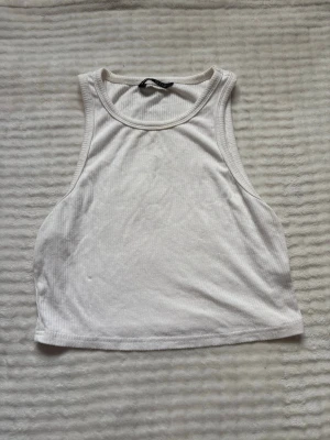 Vit ribbad croppad linnetopp - Säljer en vit ribbad croppad linnetopp med hög halsringning och ärmlös design. Perfekt till sommaren och enkel att matcha med olika stilar. Materialet är mjukt och stretchigt, vilket gör toppen bekväm att bära hela dagen.