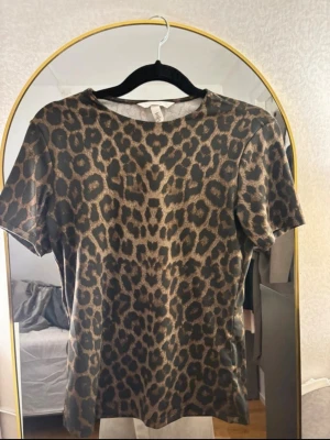 Leopardmönstrad t-shirt från H&M - Snygg leopardmönstrad t-shirt från H&M i bruna och svarta toner. T-shirten har korta ärmar och en klassisk passform. Perfekt för dig som vill sticka ut med ett djurmönster i garderoben.