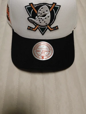 Anaheim Ducks keps Mitchell & Ness - Snygg Anaheim Ducks keps från Mitchell & Ness med broderad logga framtill och svart skärm. Kepsen är vit med svart knapp på toppen och har en broderad 'Champions' patch på sidan. Pro-Finch Fit-modell med klassisk snapback-stil. Använd en gång.