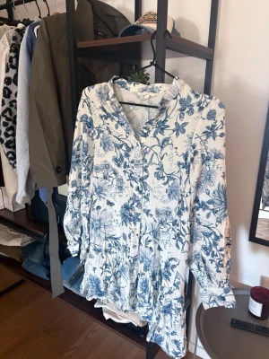 Blommig vit och blå kortklänning - Säljer en söt kortklänning med blått blommönster på vit botten. Klänningen har långa ärmar, v-ringning och volangdetaljer nedtill. Perfekt för sommarens alla tillfällen och ger en fräsch och romantisk look.