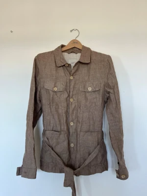 Beige linneskjorta overshirt Mango - Stilren beige overshirt från Mango i 100% linne. Skjortan har knappar framtill, två bröstfickor med lock, två stora fickor nedtill och ett knytband i midjan för en snygg siluett. Perfekt lager på lager-plagg med avslappnad känsla.