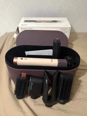 Dyson Airwrap Co-anda2x - Säljer en Dyson Airwrap Complete med flera stylingtillbehör. Setet innehåller olika borstar och cylindrar för att skapa lockar, volym och släta frisyrer. Färgen är beige med koppar- och svarta detaljer. Allt kommer i ett snyggt, lila förvaringsetui i hårdplast.