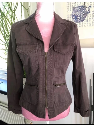 Brun figursydd jacka med dragkedja - Snygg brun jacka i figursydd modell med dragkedja framtill och fyra fickor. Jackan har klassisk krage och långa ärmar. Materialet känns som bomull och ger en avslappnad men stilren look. Färgen är grön/ brun 