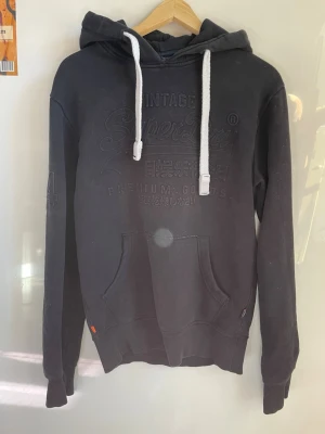 Svart hoodie från Superdry - ⚠️Inga fläckar men blev så på bilderna⚠️Säljer en svart hoodie från Superdry med broderad logga och japansk text på bröstet. Hoodien har vita dragsnören, känguruficka och ribbade muddar. Har dock ett litet hål stort som en nål på ryggen. 