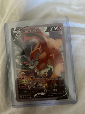 Charizard V Pokémonkort - Säljer ett Charizard V Pokémonkort med 310 HP, i färgerna röd, orange och svart. Kortet är inneslutet i ett plastfodral för extra skydd. Perfekt för samlare eller Pokémonfans som vill utöka sin samling.