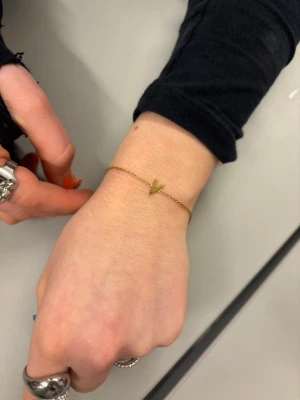 Edblad guldigt armband med V-formad detalj - Snyggt och minimalistiskt armband i guldton med en liten V-formad detalj i mitten. Tunt kedjearmband som passar perfekt för en stilren look. Justerbar längd med karbinlås. Perfekt accessoar för att lyfta din outfit.