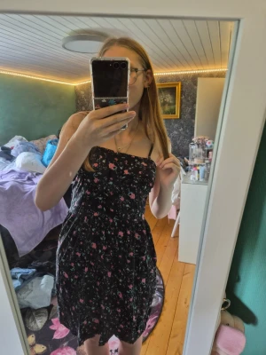 Blommig svart klänning från Vila - Supersöt svart kortklänning från Vila med rosa och grönt blommönster. Klänningen är ärmlös med smala axelband och har en lätt utställd passform. Perfekt för varma dagar och enkel att styla med sneakers eller sandaler.
