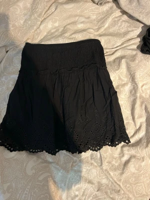 Svart kortkjol med broderi från Zara - Supersöt svart kortkjol från Zara med smock i midjan och broderade detaljer längs fållen. Kjolen har en lätt volangig form och är perfekt för dig som gillar en feminin och trendig stil.