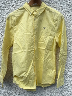Gul skjorta från Gant - Klassisk gul skjorta från Gant med button down-krage och broderad logga på bröstet. Skjortan är långärmad och tillverkad i mjuk bomull, perfekt för en stilren look.
