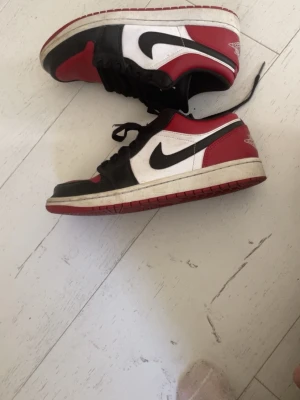 Nike Air Jordan 1 Low röd svart vit  - Snygga Nike Air Jordan 1 Low sneakers i rött köpt på footlocker, svart och vitt med klassisk swoosh på sidan som går att tvätta. Lågt skaft och ikonisk design som passar perfekt till streetwear. Skorna har en vit mellansula och röd yttersula. Materialet är en mix av läder och syntet.