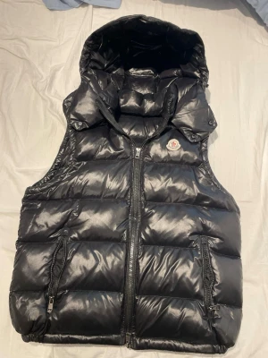 Svart dunväst från Moncler med huva - Snygg svart dunväst från Moncler med glansig finish, dragkedja framtill och två fickor med dragkedja. Västen har en stor vadderad huva och klassisk Moncler-logga på bröstet. Perfekt för lager-på-lager och streetstyle. Självklart funkar NFC-scan också.