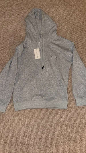 Grå hoodie med CD logga - Snygg grå hoodie med broderad logga på bröstet. Klassisk modell med huva och dragsnören, samt ribbade muddar vid ärmslut och nederkant. Perfekt för en avslappnad och trendig stil.
