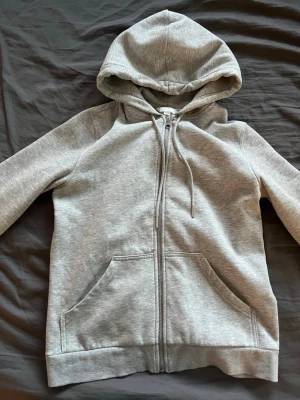 Grå zip hoodie med fickor - En stilren grå hoodie med dragkedja framtill och två stora fickor. Tröjan har huva med dragsko och ribbade muddar vid ärmslut och nederkant. Perfekt för en avslappnad look och enkel att matcha med andra plagg.