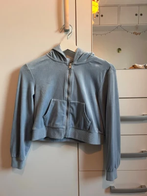 Ljusblå hoodie från bpc - Säljer en ljusblå hoodie från bpc med dragkedja framtill och känguruficka. Hoodien har en croppad passform, ribbade muddar och är gjord i mjukt velourmaterial. Perfekt för dig som gillar en chill och avslappnad stil.