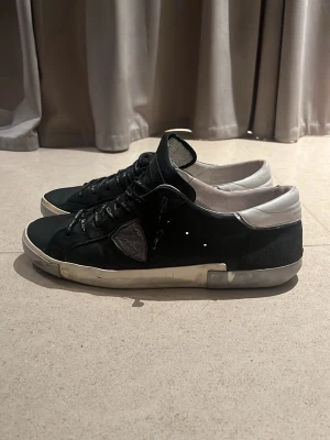 Philippe Model svarta sneakers - Svarta sneakers från Philippe Model med vita detaljer och grå logotyp på sidan. Skorna har svarta snören med vit text, beige sula och en låg siluett. Ovandelen är i läder och mocka, med vadderad häl i vitt. Perfekta för en trendig streetstyle-look.