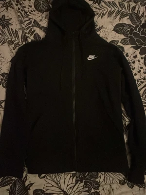 Svart Nike zip-hoodie med huva - Svart hoodie från Nike med dragkedja och huva. Klassisk design med Nike-logga i vitt på bröstet. Perfekt för en avslappnad och sportig stil. Mjuk och bekväm, långärmad modell.