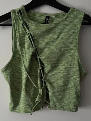 Grön croppad linnetopp med snörning från H&M - Trendig grön croppad linnetopp från H&M med diskret rutigt mönster och snygg snörningsdetalj framtill. Ärmlös modell i mjukt bomullsmaterial, perfekt för en cool och avslappnad stil eller en utekväll.