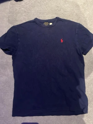 Mörkblå t-shirt från Polo Ralph Lauren - Klassisk mörkblå t-shirt från Polo Ralph Lauren med röd broderad logga på bröstet. T-shirten har rund halsringning och korta ärmar. Tillverkad i mjuk bomull för en skön känsla och enkel stil.