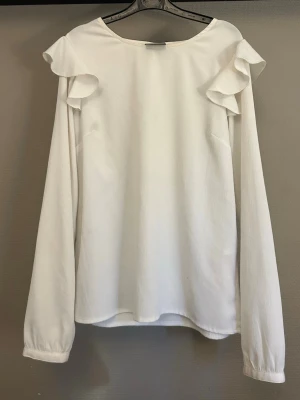 Vit blus med volangdetaljer - Stilren vit blus med långa ärmar och volangdetaljer på axlarna. Blusen har rund halsringning och är tillverkad i ett lätt, mjukt tyg som ger ett elegant intryck.