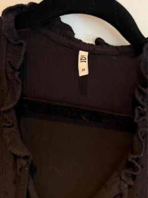 Svart blus med volang från JDY - Svart blus från JDY med korta ärmar och volangdetaljer längs ringningen. Blusen har en lätt textur i tyget och knytband vid halsen. 