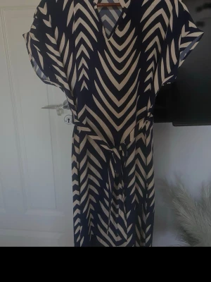 Marinblå och vit mönstrad klänning - Snygg marinblå och vit klänning med grafiskt randigt mönster och knytband i midjan. Klänningen har v-ringning och korta ärmar, perfekt för en trendig och avslappnad look.
