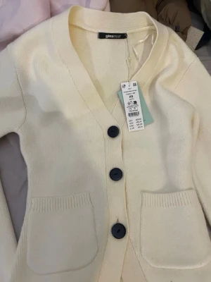 Krämvit kofta från Gina Tricot - Mysig krämvit kofta från Gina Tricot med stora svarta knappar och två framfickor. Koftan har v-ringning och ribbade muddar vid ärmslut och nederkant. 