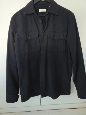 Svart overshirt med bröstfickor - Stilren svart overshirt med klassisk krage och två stora bröstfickor med lock och knapp. Långärmad modell med knäppning framtill, perfekt att bära som lager. Tyger är något tjockare, vilket ger en avslappnad men snygg look.