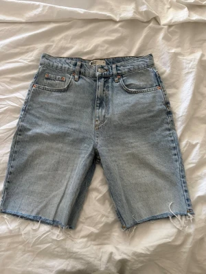 Ljusblå denimshorts med rå kant - Säljer ett par ljusblå denimshorts med hög midja och rå, fransig kant nertill. Klassisk femficksmodell med knapp och dragkedja framtill. Perfekta för en avslappnad stil.