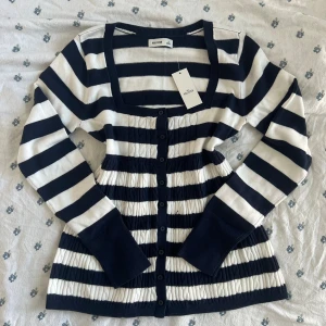 Randig babydoll kofta från Hollister, marinblå/vit - Helt ny med lappen kvar. Supersnygg babydoll kofta från Hollister i marinblått och vitt med breda ränder. Modellen har lång ärm, knappar hela vägen och markerad midja. Tillverkad i mjuk bomullsblandning med viskos. Perfekt för lager-på-lager och ger en chill men ändå stilren vibe. Måtten finns vid behöv. Storlek M / medium. Passar också till S. 