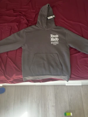 Grå Hoodrich hoodie  - Snygg grå hoodie från Hoodrich med vit texttryck på bröstet och stort tryck på ryggen. Klassisk känguruficka framtill och huva. Perfekt för dig som gillar streetwear och vill sticka ut med coola detaljer.