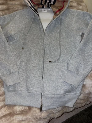 Grå hoodie från Burberry med dragkedja - Snygg grå hoodie från Burberry med dragkedja och klassiskt rutigt foder i huvan. Broderad logga på bröstet och två fickor framtill. Perfekt för en avslappnad och trendig stil. Mjukt material och ribbade muddar vid ärmslut och midja.                                                     Skicka för mer info o bilder.               Pris går o diskutera vid snabb affär!!