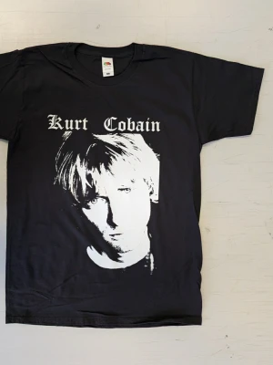 Svart Kurt Cobain t-shirt Fruit of the Loom - Svart t-shirt med stort vitt tryck av Kurt Cobain på framsidan och text i gotisk stil. Klassisk rund hals och korta ärmar. Tillverkad i mjuk bomull som känns skön mot huden. Perfekt för dig som gillar bandmerch och vill ha en ikonisk look. Helt oanvänd t-shirt med tryck Storlek: Smal 100% bomull Screentryckt