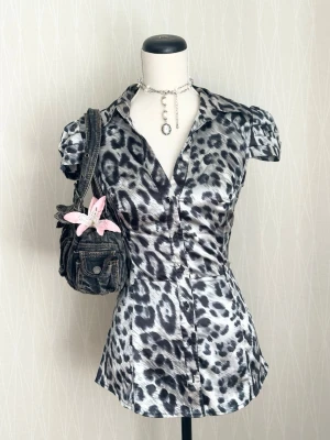 Leopardmönstrad blus med puffärm - Snygg figursydd blus med svartvitt leopardmönster och glansigt material. Blusen har korta puffärmar, krage och knäppning framtill. Perfekt för dig som vill sticka ut med en trendig look.