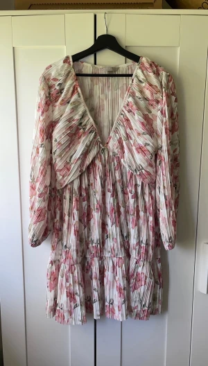 Plisserad blommig klänning - Superfin kortklänning med plisserat tyg och blommigt mönster i rosa och vitt. Klänningen har v-ringning, långa ärmar och volangdetaljer. Perfekt för midsommar etc! Från H&M i storlek XL. Helt slutsåld. 