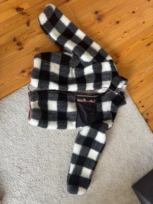 Rutig teddyjacka i svart och vitt - Mysig teddyjacka med stora svartvita rutor och dragkedja framtill. Jackan har en innerficka med dragkedja och är tillverkad i fluffigt teddy-material. Perfekt för dig som gillar en trendig och bekväm stil. Storlek 146/152 aldrig använd 