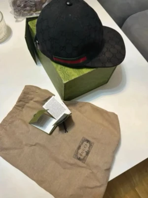 Svart keps med monogram - Snygg svart keps med klassiskt GG-monogram över hela kepsen och en grön-röd rand på sidan. Kepsen har böjd skärm och justerbar rem bak. Tillverkad i canvasmaterial och levereras med originalpåse och box.