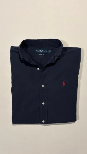 Mörkblå skjorta från Ralph Lauren - Klassisk mörkblå skjorta från Ralph Lauren med röd broderad logga på bröstet. Skjortan har knappar framtill och klassisk krage. Perfekt för en stilren look. Pris: 399