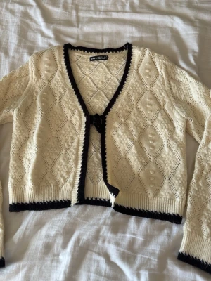 Stickad kofta från Shein i offwhite - Söt stickad kofta från Shein i offwhite med svarta kanter. Koftan har ett fint hålmönster, V-ringning och knytning framtill. Långa ärmar och ribbstickade muddar. Perfekt för lager-på-lager och en trendig look.