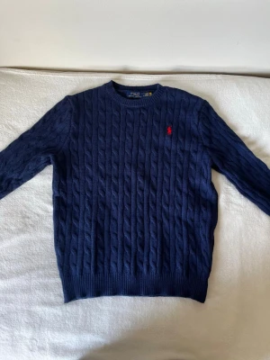 Mörkblå kabelstickad tröja Polo Ralph Lauren - Kabelstickad mörkblå tröja från Polo Ralph Lauren med rund halsringning och röd broderad logga på bröstet. Tröjan har ribbade muddar vid ärmslut och nederkant. Perfekt för dig som gillar klassisk och stilren look.