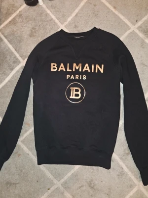 Svart Balmain Paris sweatshirt - Snygg orginal svart sweatshirt från Balmain Paris med guldig logga och tryck framtill. Tröjan har rund halsringning och långa ärmar. Perfekt för dig som vill ha en stilren och trendig look med exklusiv känsla. Trstad nångång som ny