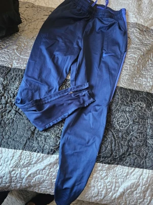 Blå Nike Dri-Fit träningsbyxor - Säljer ett par blå träningsbyxor från Nike med Dri-Fit-teknologi. Byxorna har dragkedjor vid bensluten och resår i midjan. Perfekta för träning eller sportaktiviteter. Materialet är lätt och andas bra.