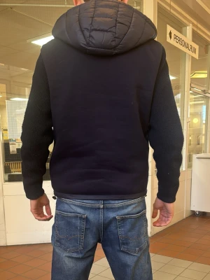 Colmar cardigan - Snygg mörkblå cardiganjacka med stickade ärmar och vadderad framsida och huva. Jackan har dragkedja framtill, två fickor med dragkedja och en patch på ärmen. Perfekt för lager-på-lager och streetwear-stil.