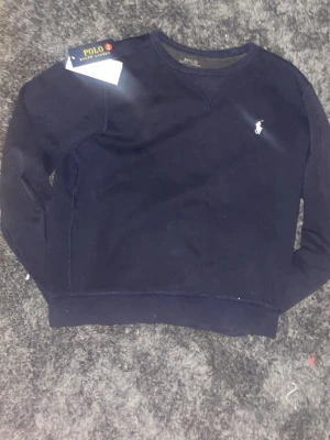 Mörkblå sweatshirt från Polo Ralph Lauren - Mörkblå sweatshirt från Polo Ralph Lauren med klassisk rund hals och vit broderad logga på bröstet. Tröjan har långa ärmar och ribbade muddar vid ärmslut och nederkant. Perfekt för en avslappnad och stilren look.