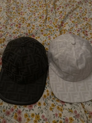 Fendi kepsar svart och vit FF-mönster - Två kepsar från Fendi, en svart och en vit, båda med det klassiska FF-logomönstret över hela kepsen. Kepsarna har böjd skärm och justerbar baksida. Materialet ser ut att vara slitstark bomull eller bomullsblandning. Perfekt för dig som gillar exklusiva accessoarer. 700FÖR EN!!!! FINNS OCKSÅ EN MÖRKBLÅ!!!!!