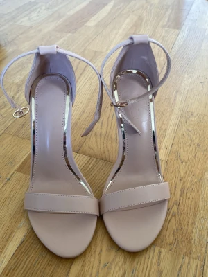 Nude pumps med ankelrem - Snygga nude-färgade pumps med öppen tå och smal ankelrem med spänne. Skorna har en enkel och stilren design med hög klack och slät yta i syntetmaterial. Perfekta för att lyfta vilken outfit som helst. Klacken är 10cm. aldrig använda 