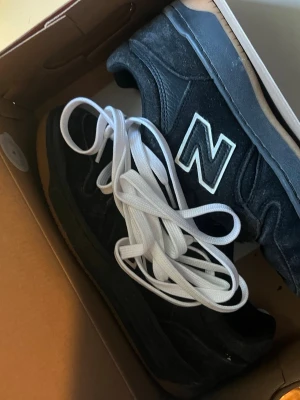 Svarta New Balance sneakers i mocka - Säljer ett par svarta New Balance sneakers i mocka och läder med klassisk vit N-logga på sidan. Skorna har svart sula med gummidetaljer och levereras med både svarta och vita skosnören. Perfekta för dig som gillar stilrena och sportiga sneakers. Original box och extra snöre med kommer också 