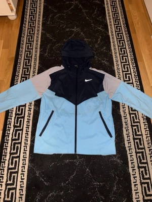 Nike miler vindjacka blå (slut i alla butiker) - Nike vindjacka med huva i blått, mörkblå och grått. Jackan har dragkedja framtill, två snedställda fickor med dragkedja och meshfoder. Perfekt för löpning eller när du vill ha en sportig look. Lätt material och snygg design med Nike-logga på bröstet.
