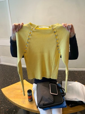 Gul ribbad topp med knappar från Zara - Trendig gul långärmad topp från Zara med ribbad struktur och dekorativa knappar längs axlarna. Toppen har en rund halsringning och är figurnära i modellen. Perfekt för dig som vill sticka ut med färg och detaljer. Jätte bra skick, aldrig använd, väldigt populär och säljs inte längre. Storlek M men liten i storleken så passar xs-s